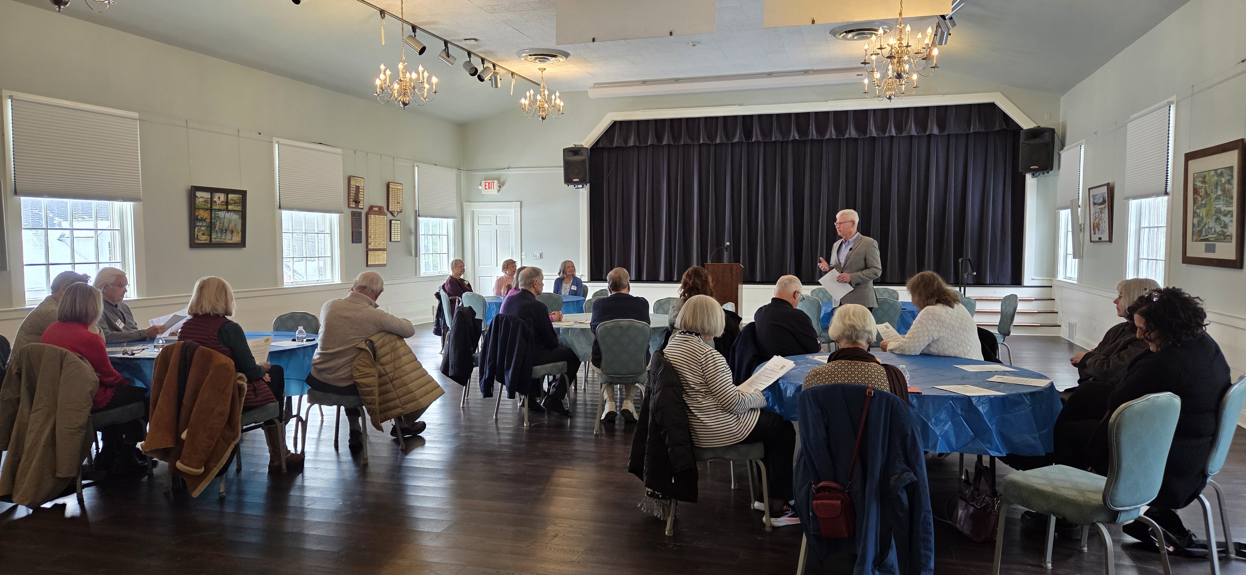 a room where people are sitting around round tables enjoying lunch and listenting to someone speaking