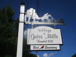 A white metal pole containing a black sillouette of trees and a man riding a horse. A white sign with black letters hangs beneath the sillouett and contains the words Village of Gates Mills Bird Sancutary