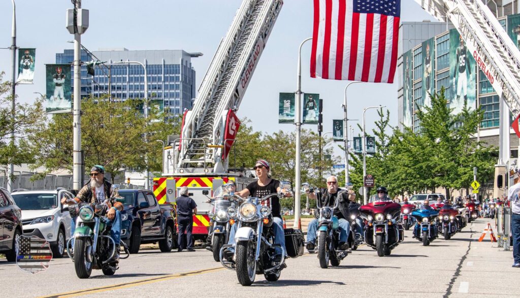 several motorcycles driving in formation two by two under the red, white and blue United States Flag