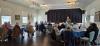 a room where people are sitting around round tables enjoying lunch and listenting to someone speaking