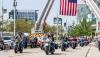 several motorcycles driving in formation two by two under the red, white and blue United States Flag